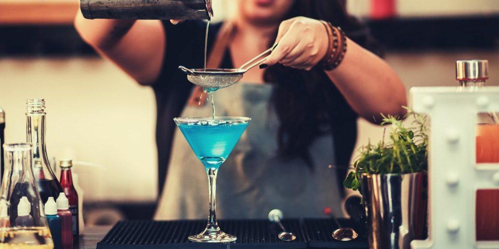 A woman straining a bright blue low alcohol cocktail into a martini glass
