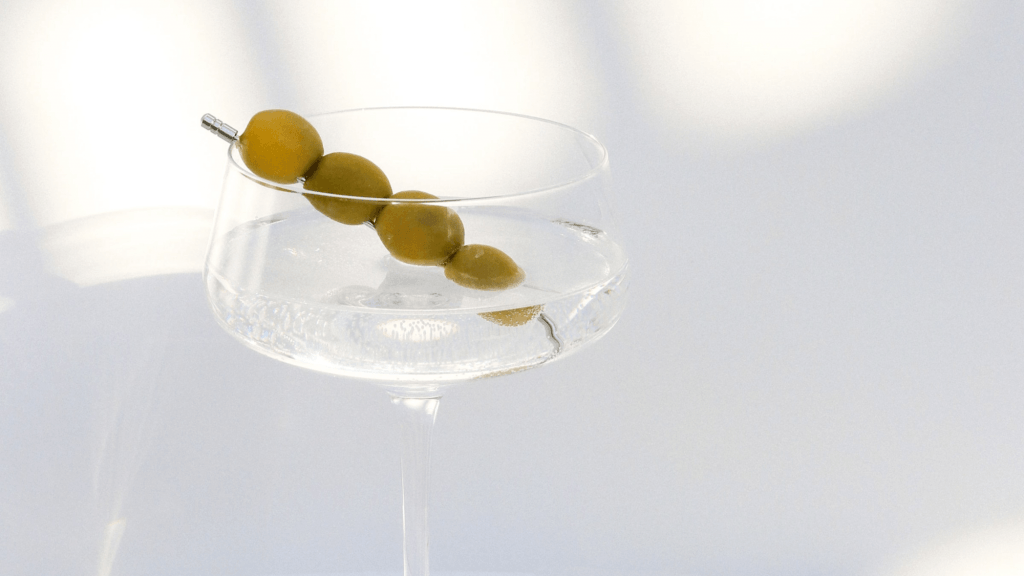 Close up of a Gin Martini garnished with green olives on a garnish pick against a sun-dappled white background