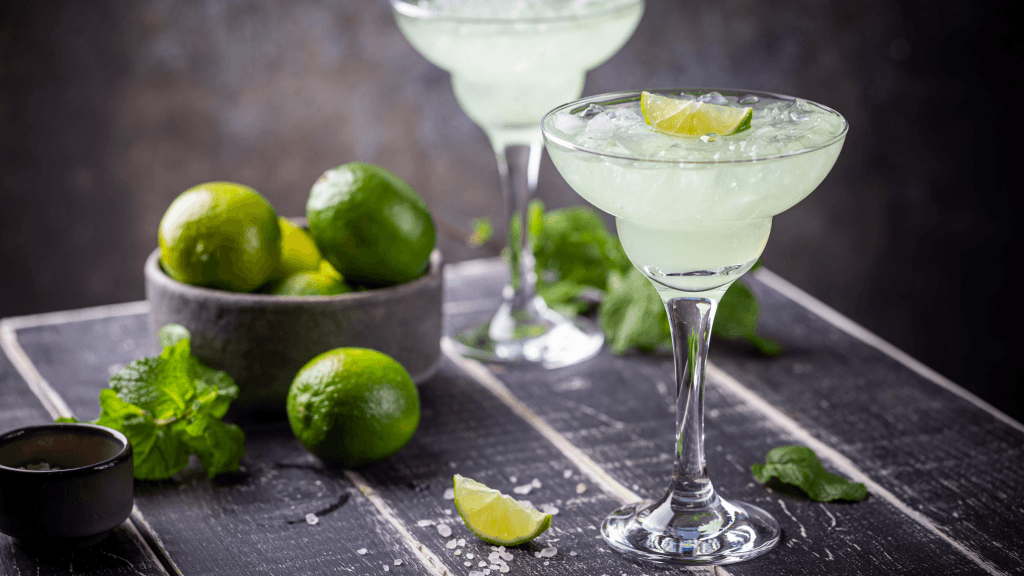 Close up of a pair of Virgin Margaritas on a grey surface, against a grey backdrop, with a bowl of limes and scattered salt around it
