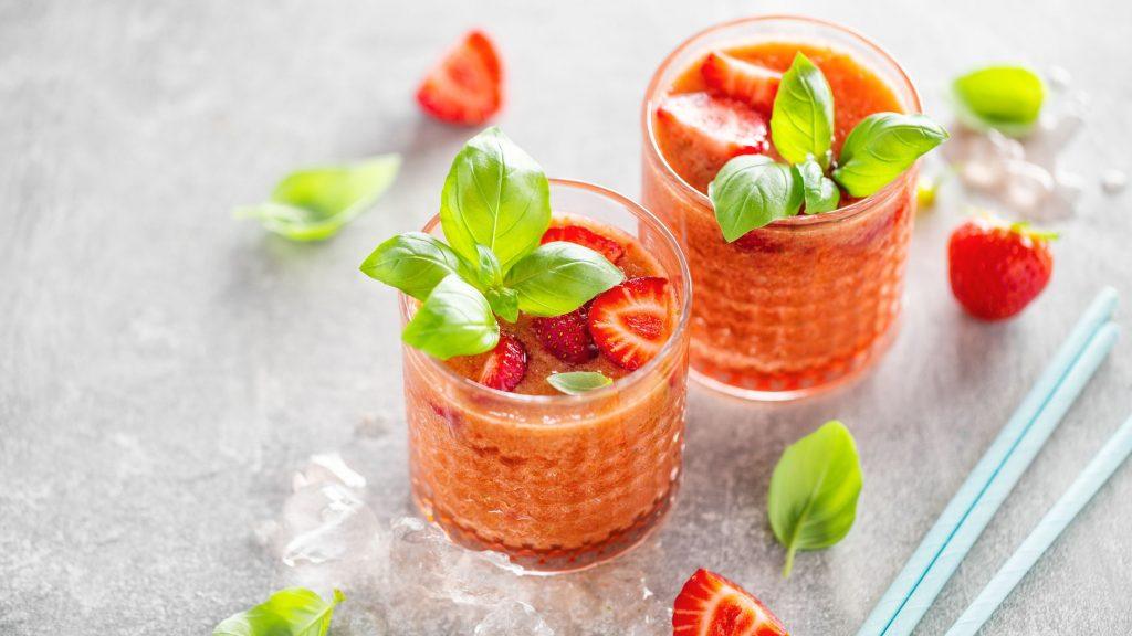 Close up top view of two Strawberry Basil cocktails in textured rocks glasses on a light grey surface, surrounded by scattered basil leaves and strawberry wedges