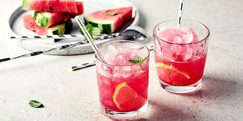 Close up front view of two Watermelon Gin cocktails in rocks glasses on a speckled marble surface, with a white serving platter laden with fresh slices of watermelon in the background