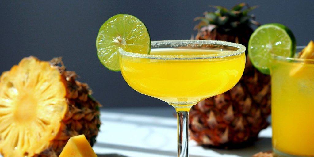 Close up front view of a Buena Vista cocktail in a Margarita glass with lime wheel, with fresh pineapples in the background