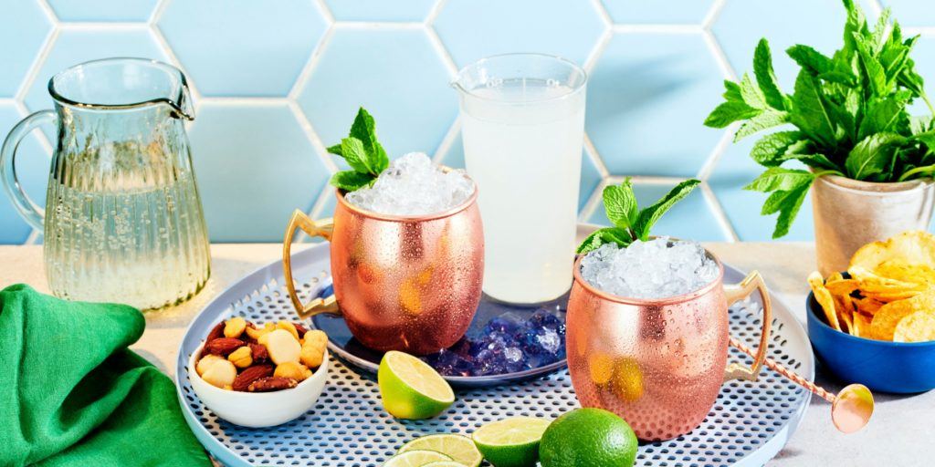 Front view of two Virgin Moscow Mules in copper mugs, on a speckled blue serving platter, surrounded by sliced limes and assorted snacks, including nuts and chips, against a light blue backdrop