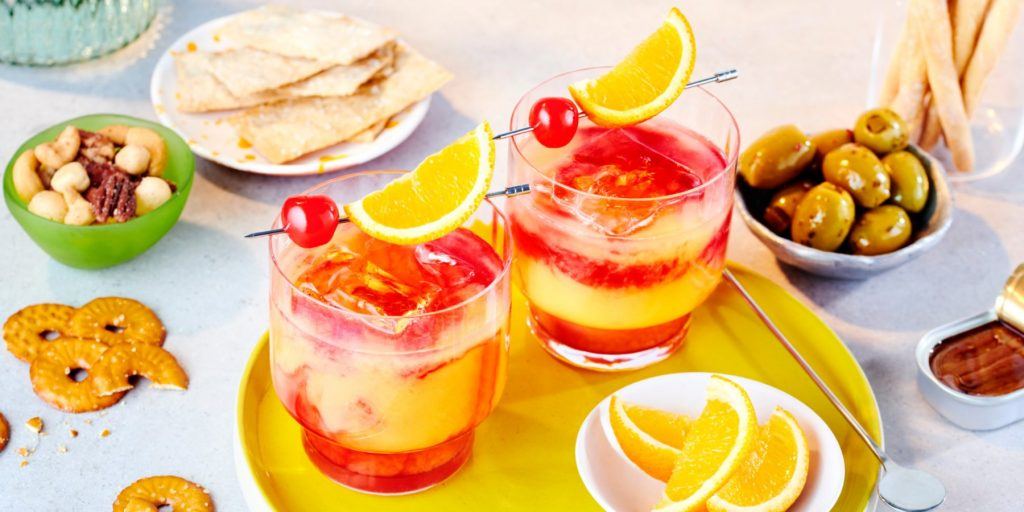 Top shot of two Tequila Sunrise cocktails garnished with cherries and orange wedges, set on a yellow serving platter surrounded by assorted snacks like pretzels and green olives