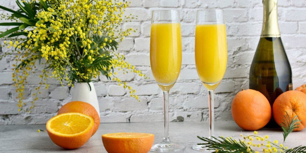 Front view of a pair of Mimosa cocktails against a white brick wall with sliced oranges alongside it, and a bottle of bubbly and a white fase with yellow flowers in the background