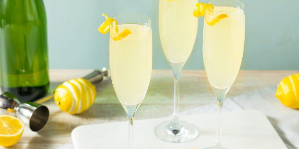 Front view of a trio of French 75 cocktails in Champagne flutes with twirly lemon twist garnishes againts a light blue backdrop