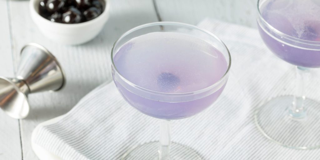 Close up top view of a pair of violet-hued Aviation Cocktails on a white surface with a bowl of black olives and a cocktail jigger visible to the side