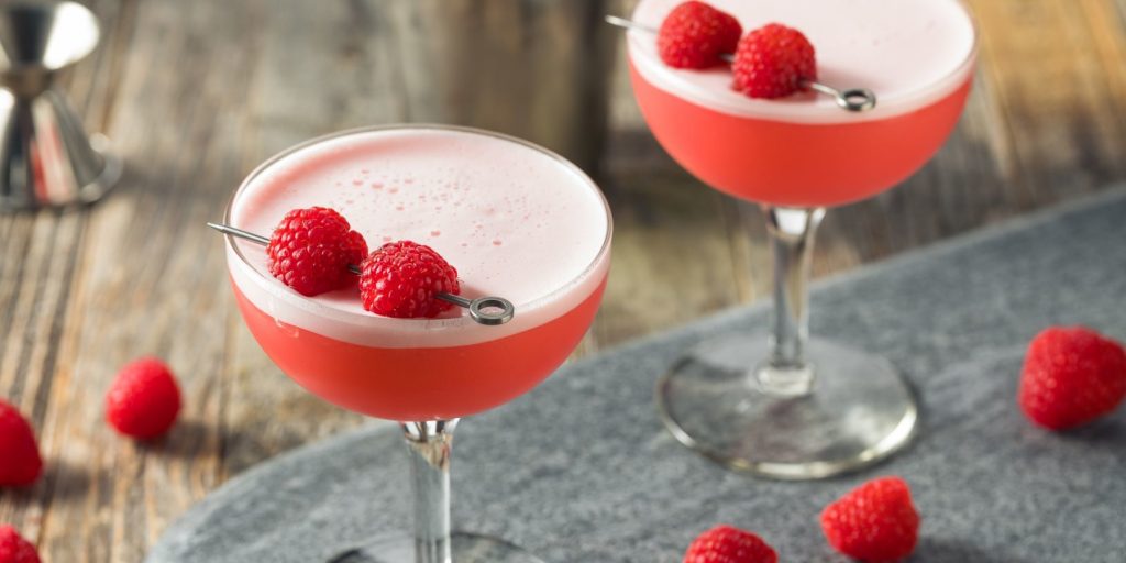 Top view of a pair of frothy Clover Club cocktails garnished with fresh raspberries on a grey surface scattered with furhter raspberries