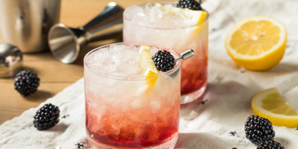 Front view of a refreshing pair of Gin Bramble Cocktails garnished with fresh berries and sliced lemon, on a white napkin, surrounded by further fresh berries and sliced lemons, with a metal cocktail shaker and jigger in the background