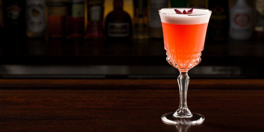 Close up front view of a French Rose cocktail garnished with rose petals, on a dark wooden surface against a dark backdrop