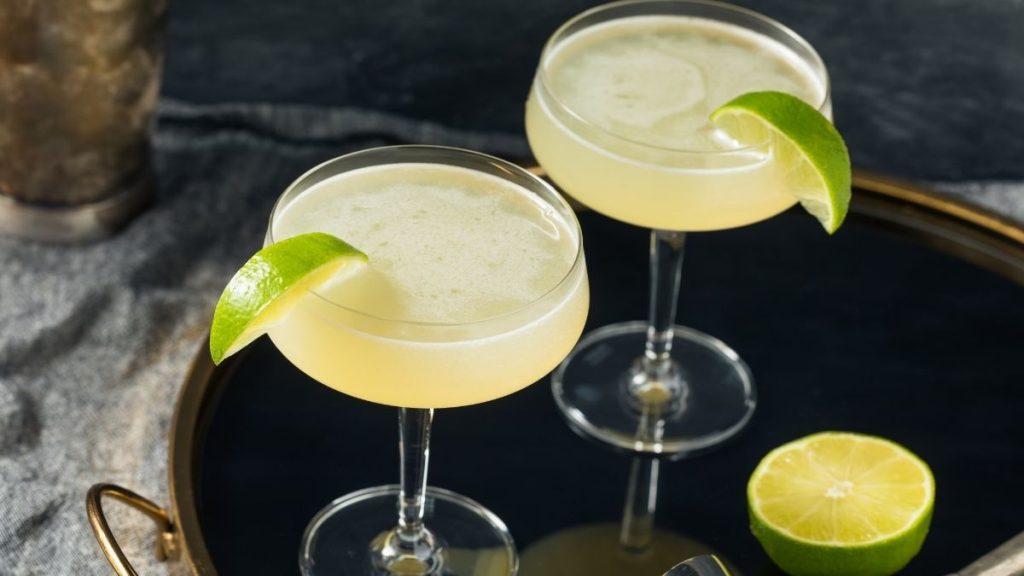 Close up to view of two French Pearl Cocktails in coupe glass, garnished with lime wedges, on a dark blue serving platter rimmend in gold