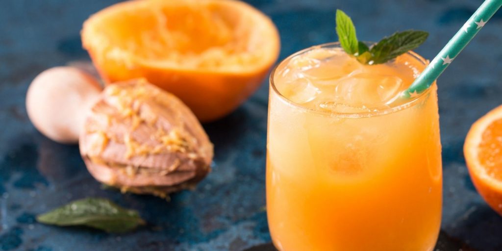 Close-up of a zesty & lovely Orange Blossom Cocktail garnished with a sprig of mint, set on a blue surface with a hand-held wooden citrus juicer and juiced orange in the background