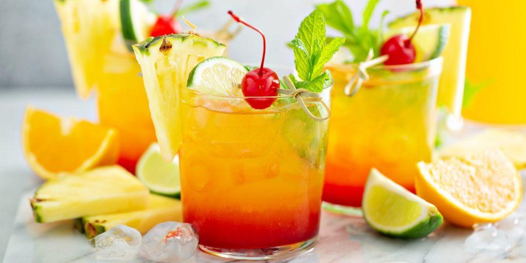 Front view of three No Tequila Sunrise Mocktails garnished with cherries and fresh lime, among fresh sliced pineapples, lime and oranges
