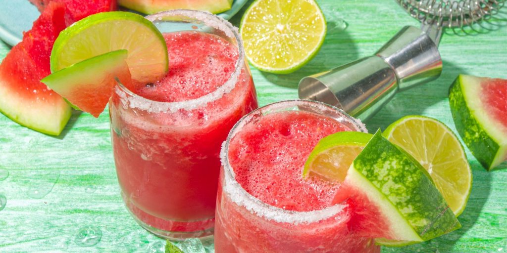 Closeup top view of two Watermelon Tequila cocktails in tumblers garnished with fresh watermelon and lime, set on a green surface with cut limes and fresh watermelon around it