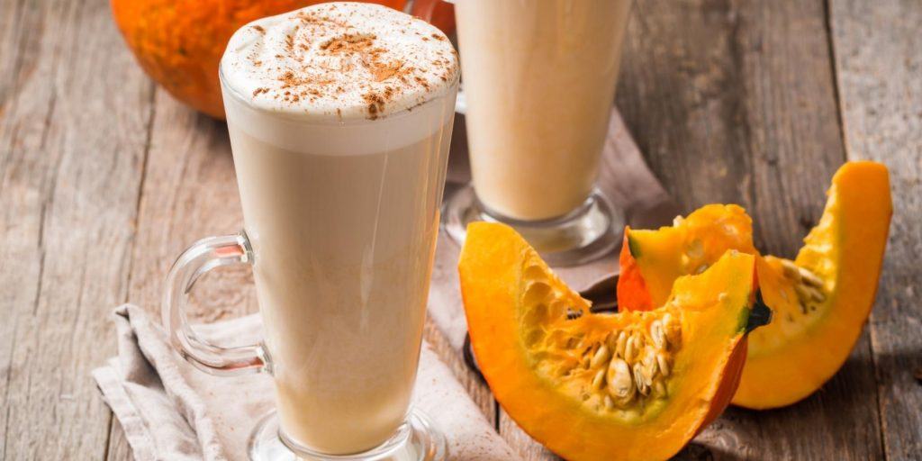 Close up front view of a pair of Hot Pumpkin Pie cocktails garnished with cinnamon, on a wooden surface, surrounded by cut-up pumpkin slices