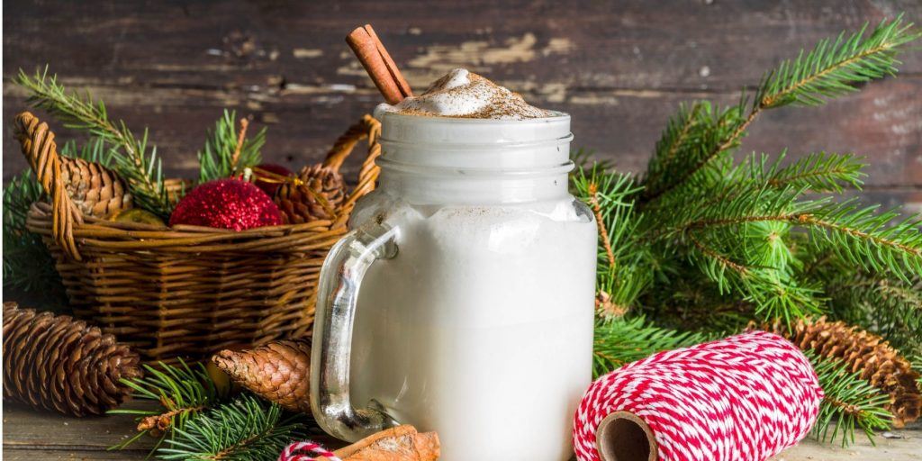 Front view of a creamy Tom and Jerry Cocktail in a mason-jar style mug, garnished with cinnamon, and surrounded by Christmas decor elements 