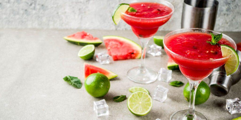 Close up front view of two Watermelon Margaritas garnished with lime, set on a grey surface with fresh limes, and slices of watermelon in view