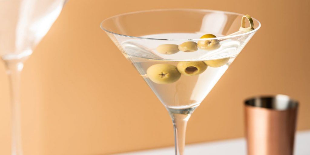 Front view of a Vodka Martini garnished with green olives against a light orange backdrop, set on a white surface