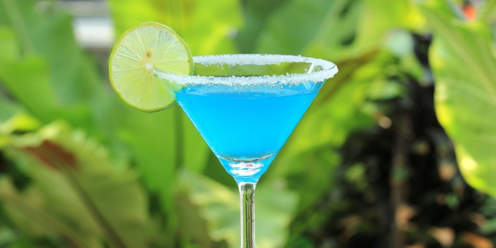 Close up front view of Blue Lagoon Margarita cocktail in front of a tropical foliage backdrop