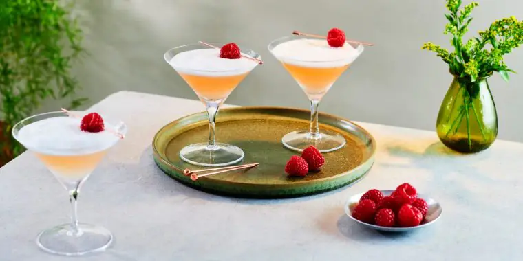 Two frothy pink French Martinis garnished with fresh raspberries placed on a silver serving platter on a table covered in a white tablecloth with a bowl of fresh raspberries and one more French Martini in the foreground