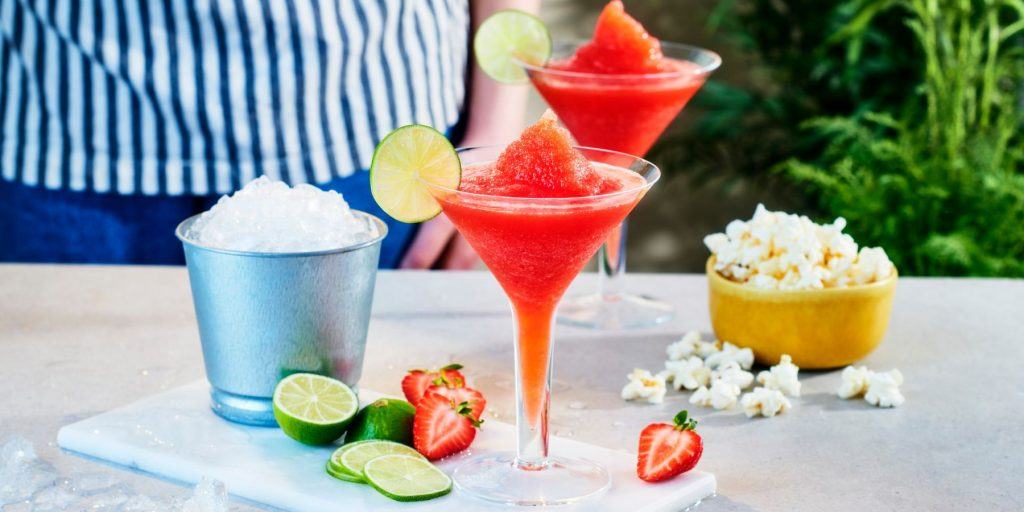 Front view of two glasses of Frozen Strawberry Daiquiri presented on a white marble surface, surrounded by a bowl of ice and a bowl of popcorn