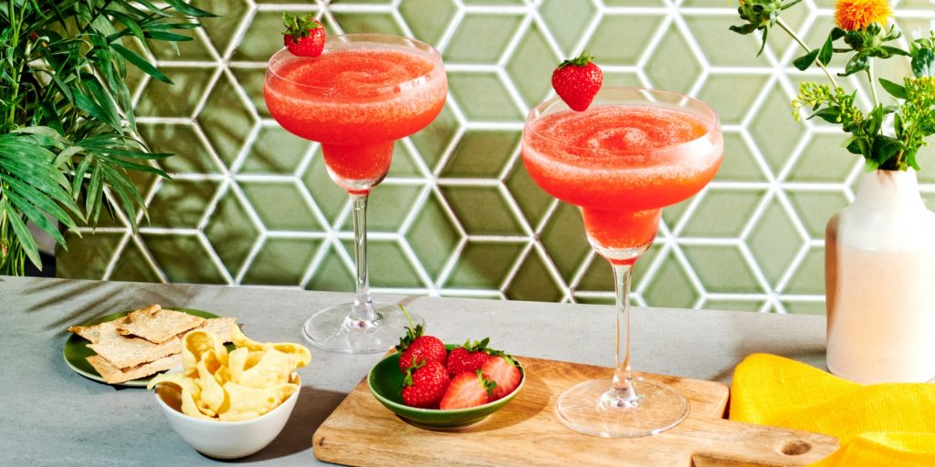 Close up front view of a pair of Virgin Strawberry Daiquiris on a grey surface, against a green tiled backdrop, accompanied by a selection of snacks, including crackers and berries in bowls