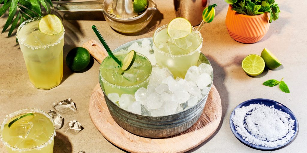 Top image of refreshing and citrusy Lime Margarita Cocktails in a bucket with ice, surrounded by more drinks and assorted garnishes, including sliced lime and a saucer of salt