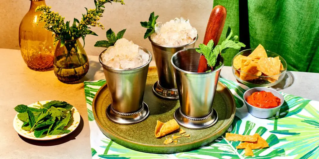 Front view of a trio of R=refreshing Mint Julep Cocktails served in copper cups on a green serving platter, surrounded by assorted snacks including chips and salsa, with a green curtain and vases with green foilage in the background
