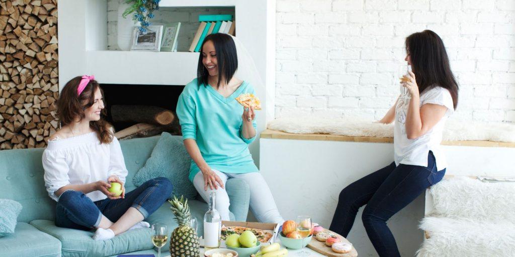 Three female friends sitting on a couch in comfortable clothing enjoying snacks and having a planning discussion for a Hen Do