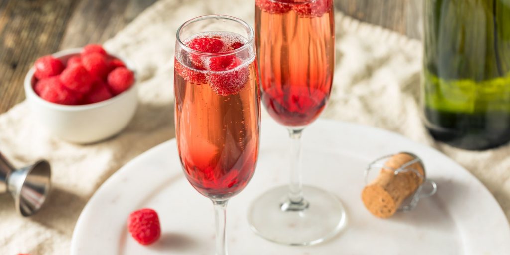 Close up top view of a pair of Kir Royale cocktails garnished with fresh raspberries, on a white serving plate on a white cloth napkin on a wooden table, with a bowl of fresh raspberries and a bottle of bubbly in view to the sides