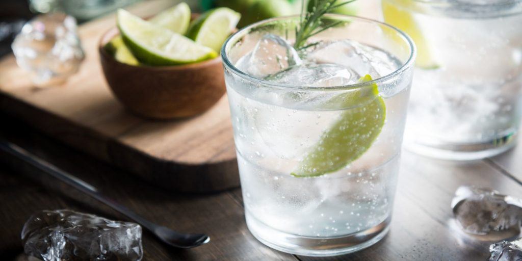 Close up front view of a pair of refreshing Gin Ricky cockftails garnished with fresh lime wedges, on a dark wood surface with a wooden cutting board with assorted garnishes in the background