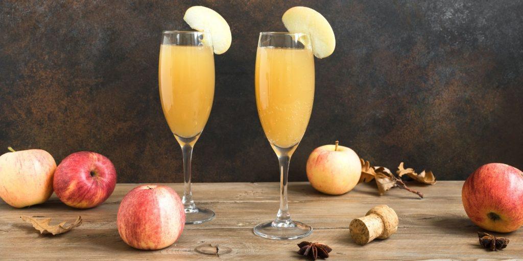 Front view of a pair of Apple Cider Mimosas garnished with slices of fresh apple, on a wooden surface with fresh apples scattered around, against a grey backdrop