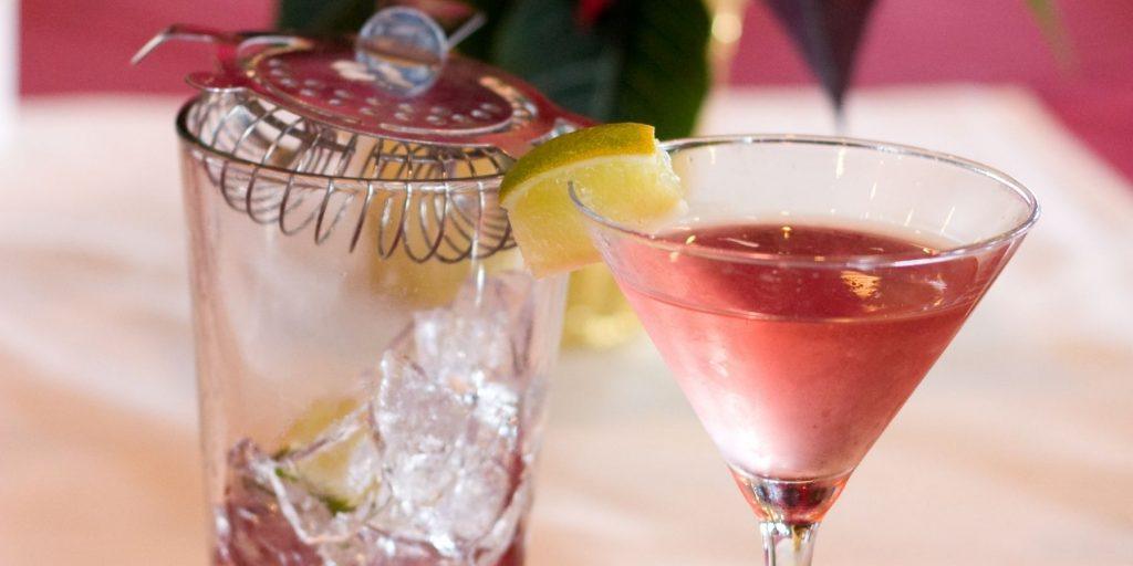 Close up of a pink Poinsettia Cocktail garnished with a lime wedge, alongside a cocktail glass with ice with a metal strainer atop it