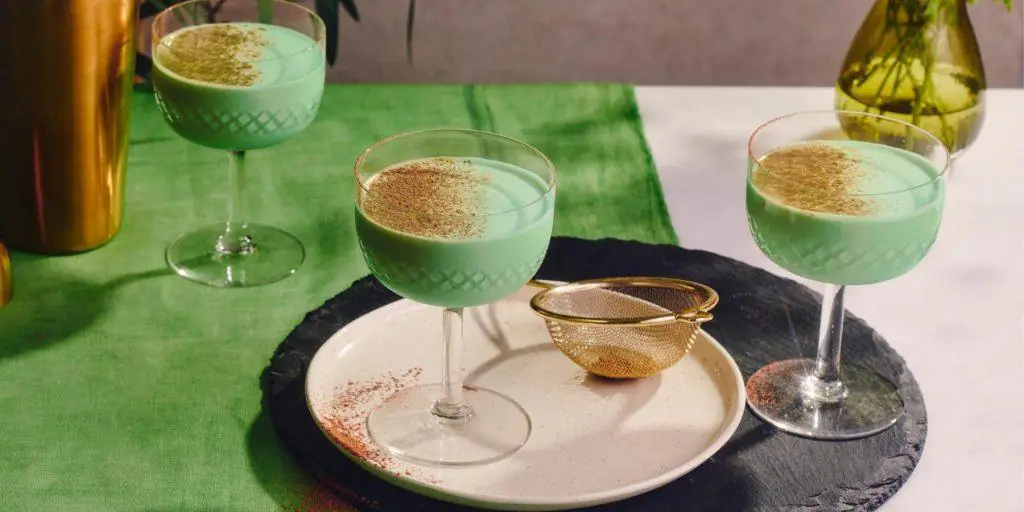 Creamy minty green Grasshopper Cocktails presented on a table covered in a green table cloth, party posed on a white serving platter on top of a slate grey placemat