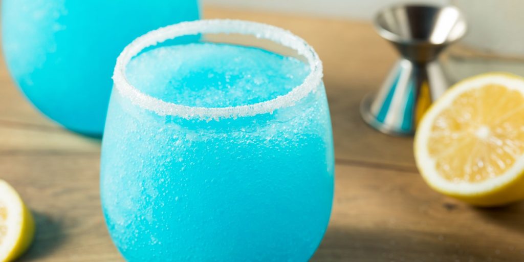 Close-up of a pair of bright blue Jack Frost cocktails rimmed with sugar, set on a wooden surface, surrounded by sliced lemons