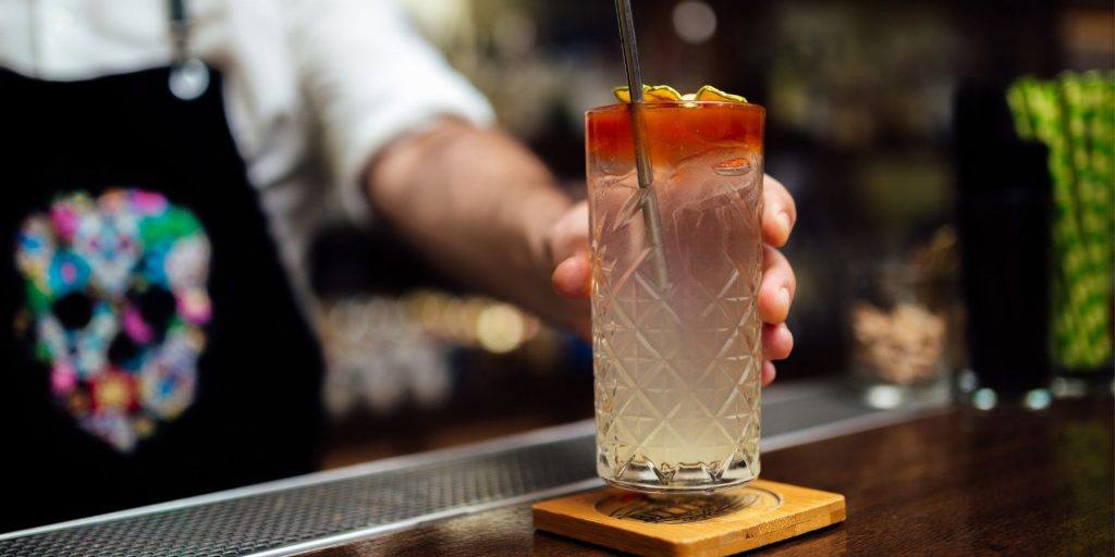 Close up of a bartender's hand serving a Bitter Collins in a Collins glass