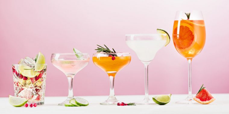 Front view of five cocktails in different glasses against a pale pink backdrop