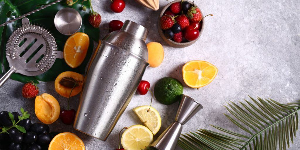 Top view of a flat lay of a cocktail shaker, strainer and other tools among a selection of seasonal cocktail ingredients, including apricots and berries, on a light grey granite surface