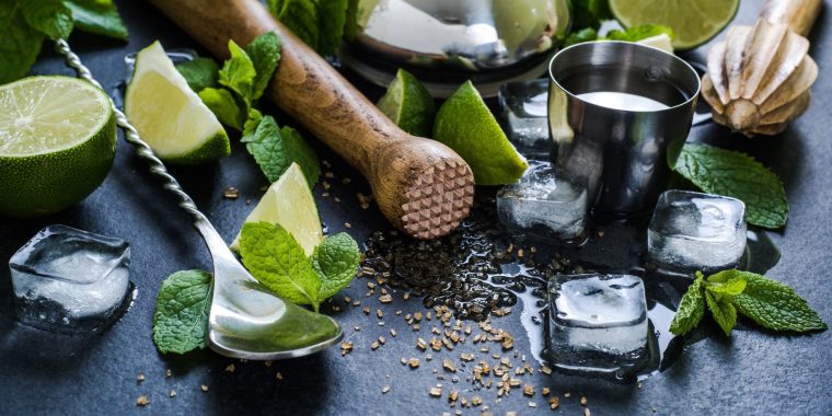 Close up top shot of a wooden cocktail mudler and metal cocktail spoon on a black surface, surrounded by sliced limes, fresh mint, and ice cubes