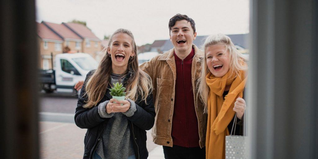 Three friends arriving at a birthday party, holding gifts, and being welcomed at the door by the host