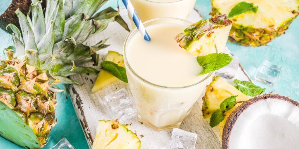 Top view of a pair of Virgin Pina Coladas garnished with fresh pineapple, amidst fresh pineapple and coconut on a light blue surface