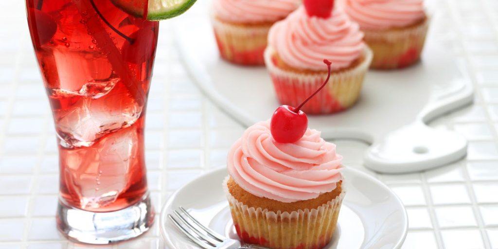 Close up front view of a Shirley Temple Mocktail served alongside cupcakes with pink icing, topped with cherries