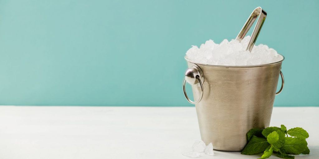 Front view of a silver ice bucket filled with ice, on a white surface against a light blue backdrop