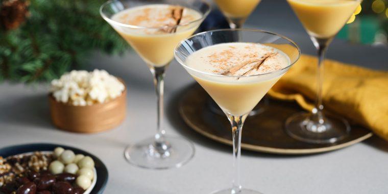 A trio of indulgent Eggnog Martini cocktails presented in long-stemmed Martini glasses, surrounded by snack bowls on a light grey surface