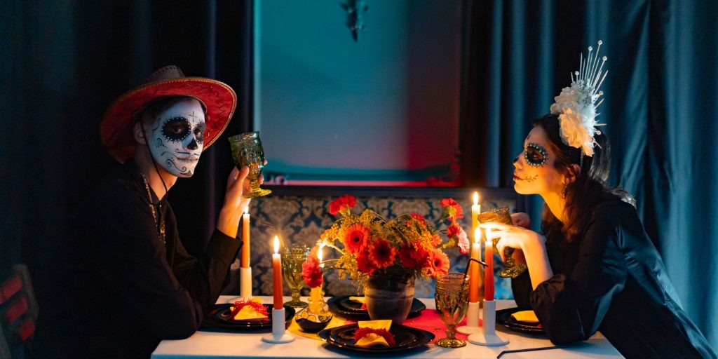 A man and woman sitting at a candlelit table wearing costumes for a Mexican Day of the Dead celebration, including sugar skull makeup