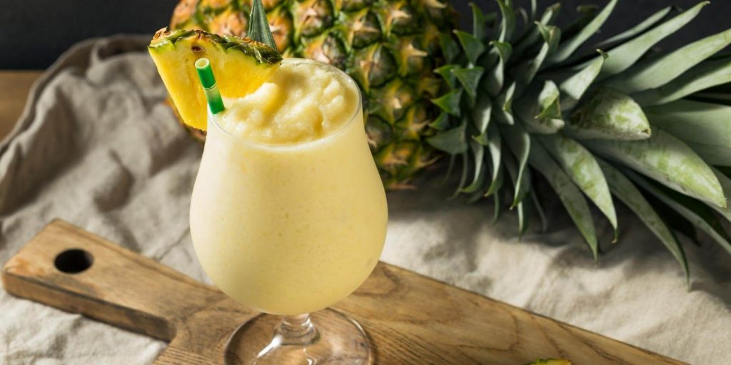 Close up of a creamy Frozen Pina Colada cocktail with pineapple garnish set on a wooden board with a fresh pienapple in the background
