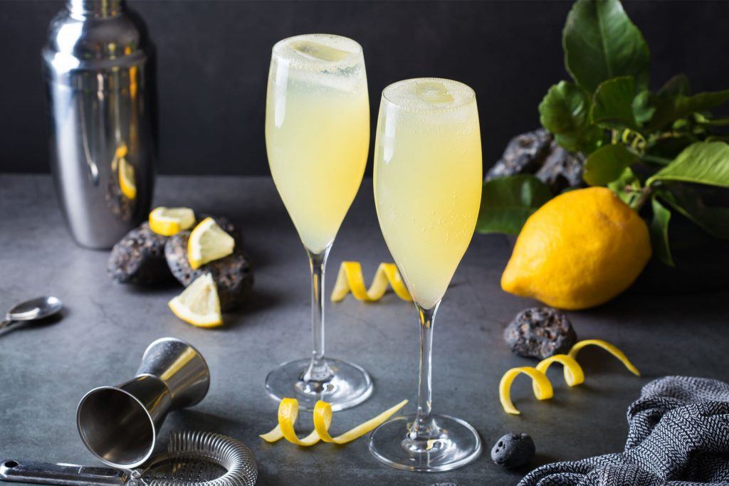 Close up of two glasses of French 75 cocktail in Champagne flutes, presented on a slate grey surface, surrounded by lemon peels, a fresh lemon, a cocktail shaker, jigger & other cocktail making tools