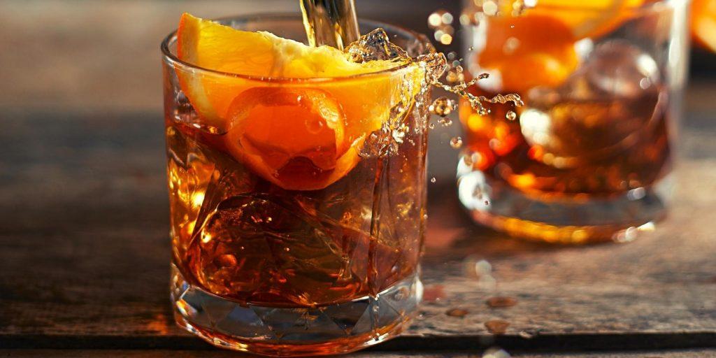 Close up of two Old Fashioned cocktails made with Japanese whiskey, a splash of liquid escaping the rim of one glass. Both glasses are presented on a rustic wooden surface.