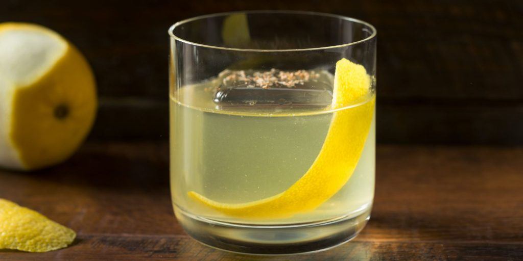 Clarified Milk Punch in a clear tumbler, on a wooden surface, garnished with a lemon peel, with a lemon in the background