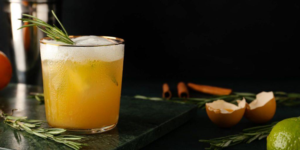 Close-up of a Whiskey Sour Cocktail Recipe on a dark surface, surrounded by cracked egg shells and assorted drinks garnishes